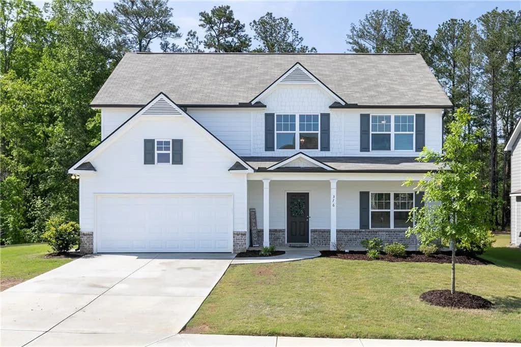 Traditional two-story home with HardiPlank siding, brick watertable foundation, covered front porch, black shutters, and an attached 2-car garage. Landscaped beds and mature tree line in the background.