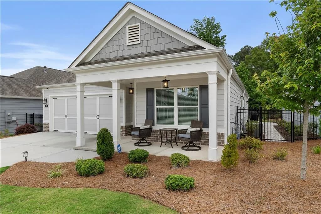 Craftsman-style exterior featuring a covered porch with white columns, a two-car garage with white doors, gray siding and shingle accents, dark shutters, and a landscaped front yard with pine straw and plantings