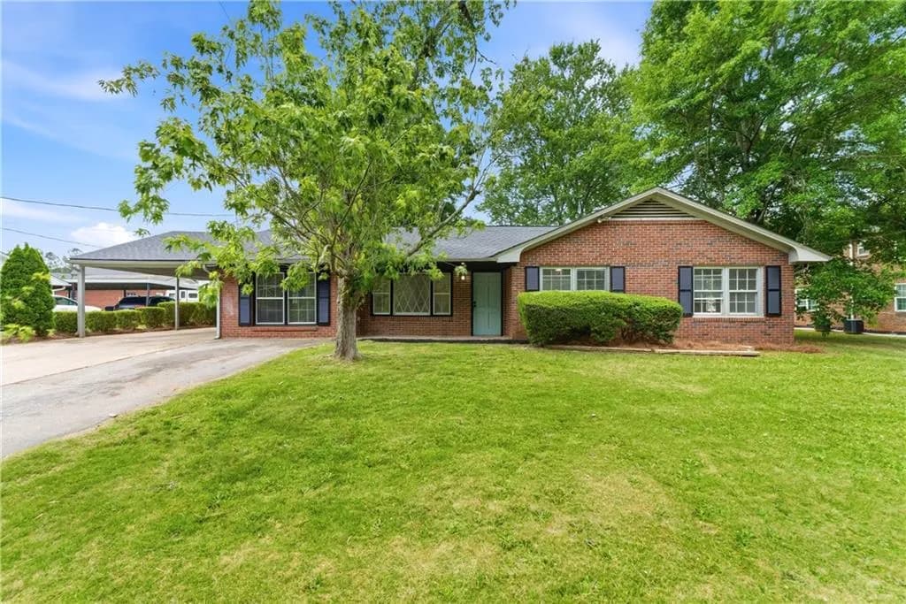 Brick exterior residence featuring a covered carport, pitched roof, front entry door, front lawn, and mature trees